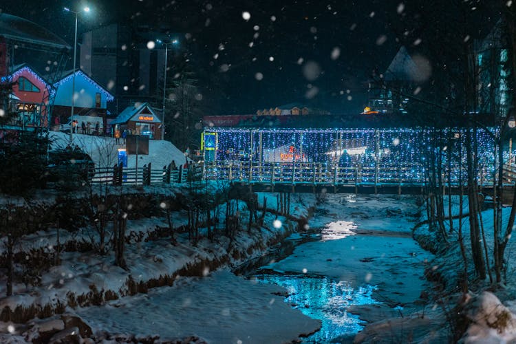 Bridge With String Lights Above A Snow Covered River At Night Time