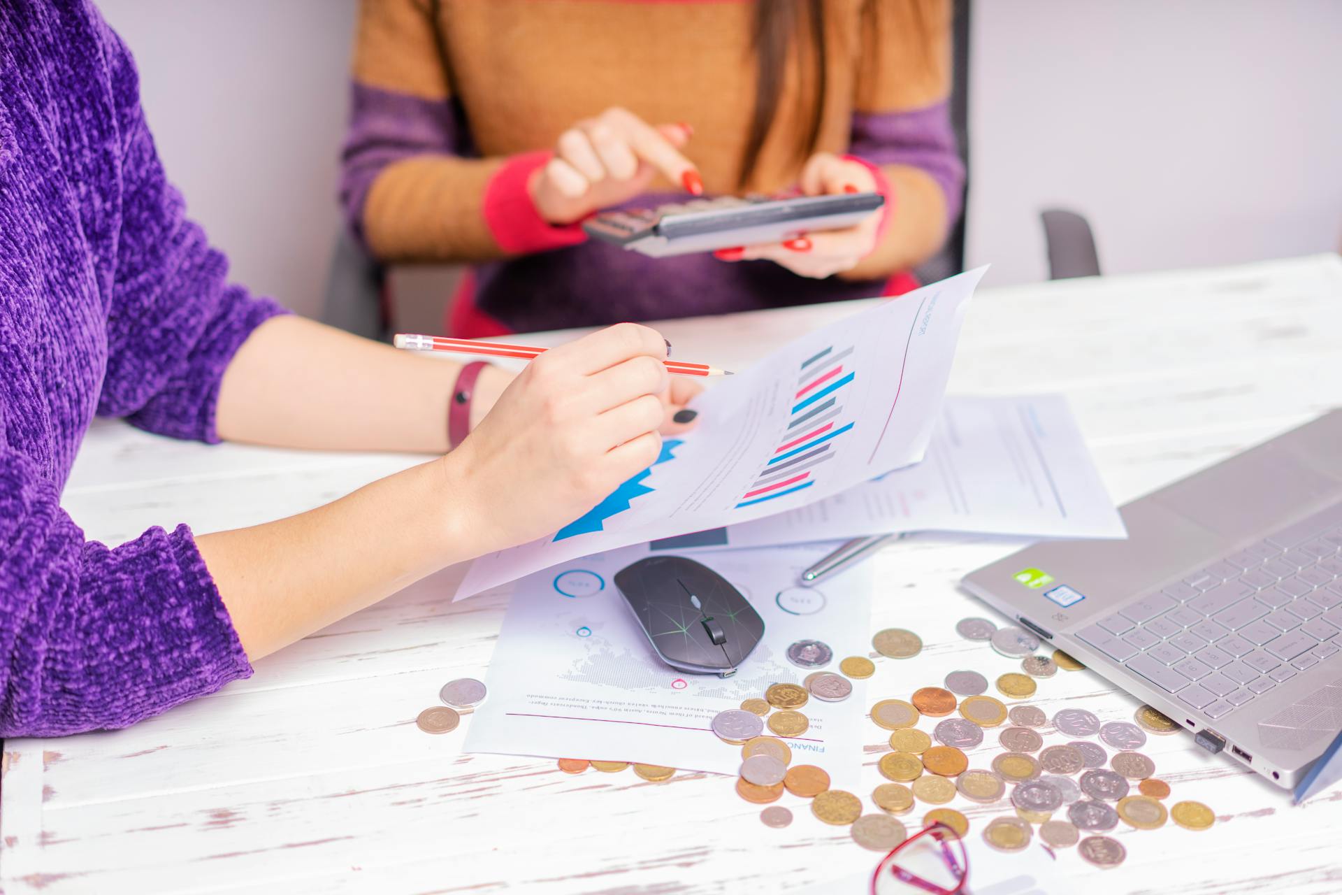 Two women analyzing financial charts with a laptop and coins on desk in an office setting.