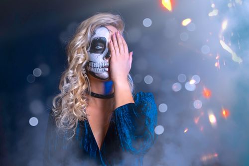 Free A blonde woman with skull face paint covers one eye, celebrating Halloween in a blue dress. Stock Photo