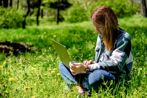 Free A woman sitting on grass using a laptop, enjoying remote work in a sunny park setting. Stock Photo