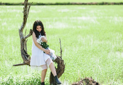 一名年轻女子身穿白色连衣裙，坐在田野里充满活力的绿色树干上，手里拿着鲜花