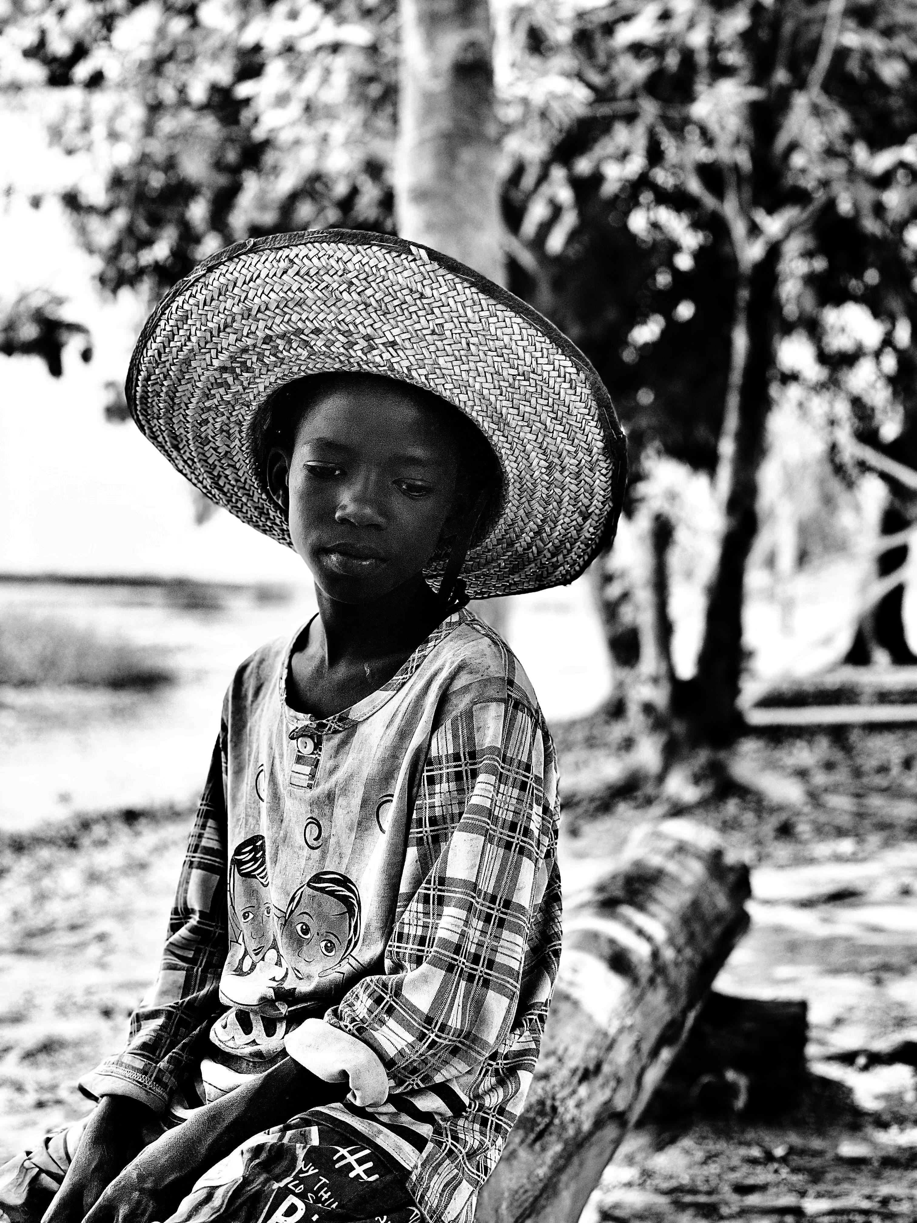 A monochrome image capturing a thoughtful child in a large straw hat, sitting outdoors.