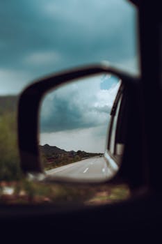 Reflection of a scenic road in a car side mirror with mountains and cloudy sky.