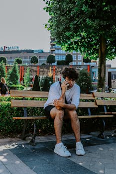 Young man sits on a wooden bench, engaged in a phone call with urban backdrop.