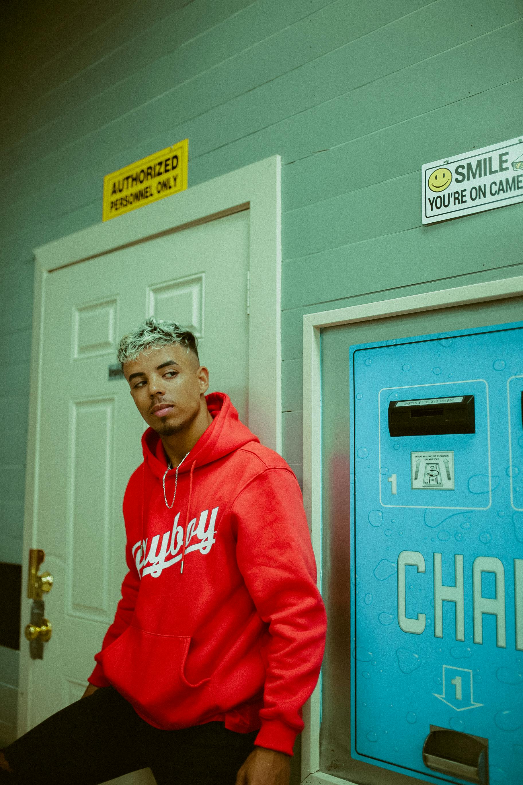 Casual portrait of a young man in a red hoodie standing next to a vending machine, indoors.