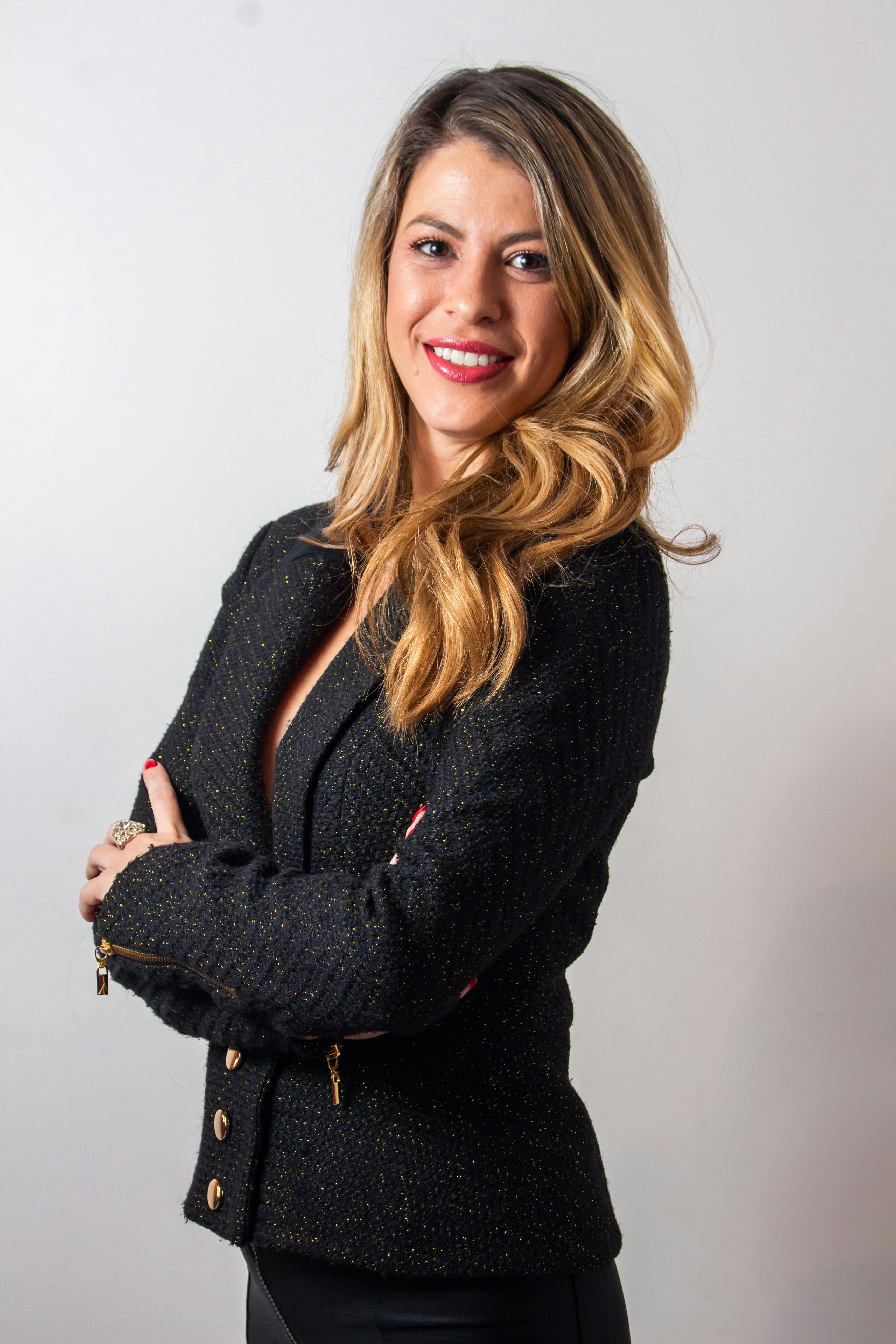 Portrait of a confident woman with blond hair in a black blazer, smiling warmly against a plain background.