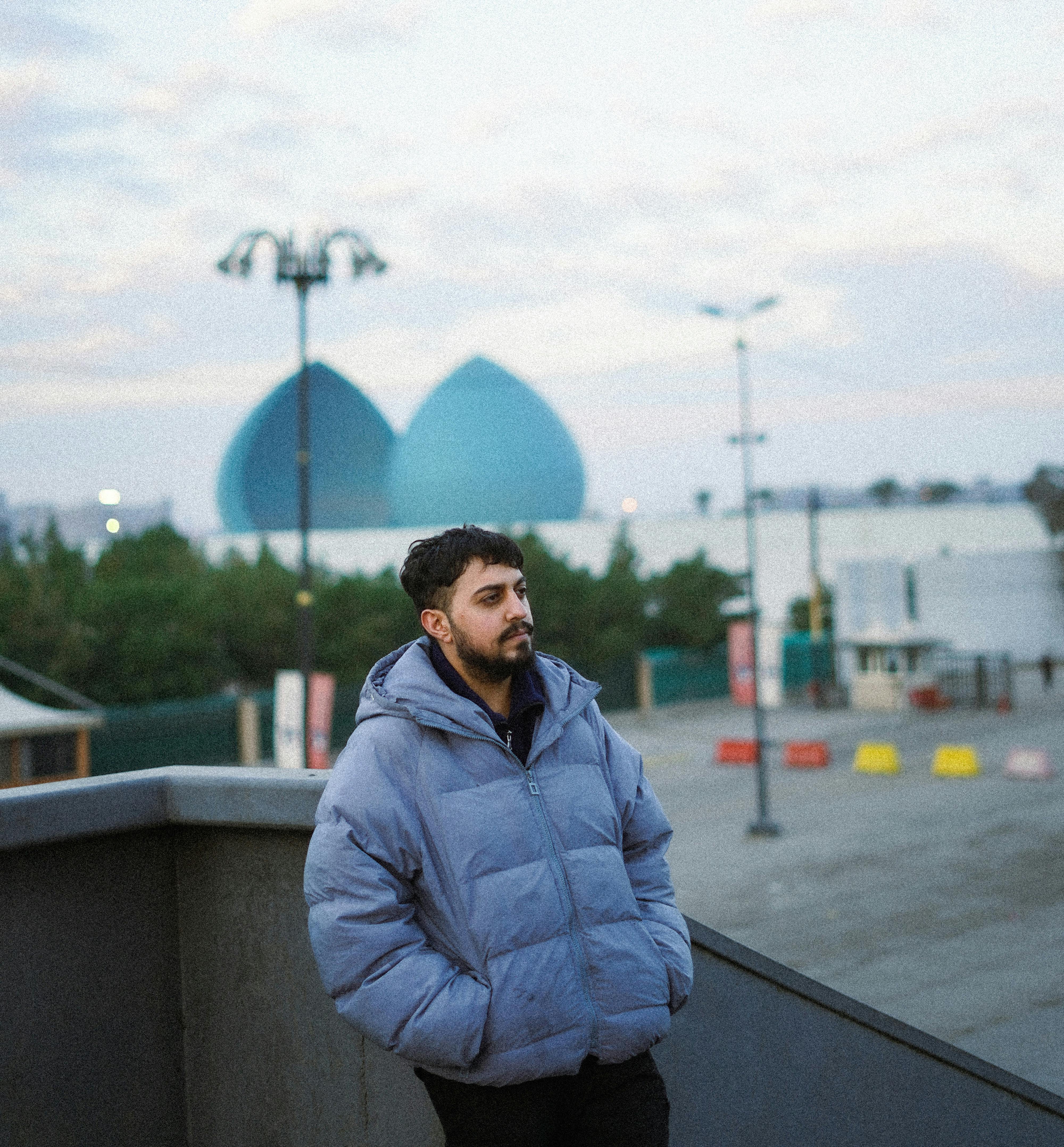 A man in a blue jacket stands on a city street with a unique dome structure behind him.