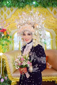 Beautiful bride in traditional attire holding a floral bouquet with an ornate crown inside a decorated indoor setting.
