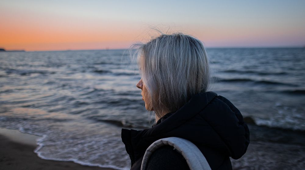 Woman in a dark hooded jacket looking out over the sea at sunset on a cool evening
