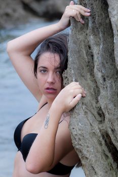 Woman in a black bikini posing confidently by seaside rocks, exuding strength and beauty.