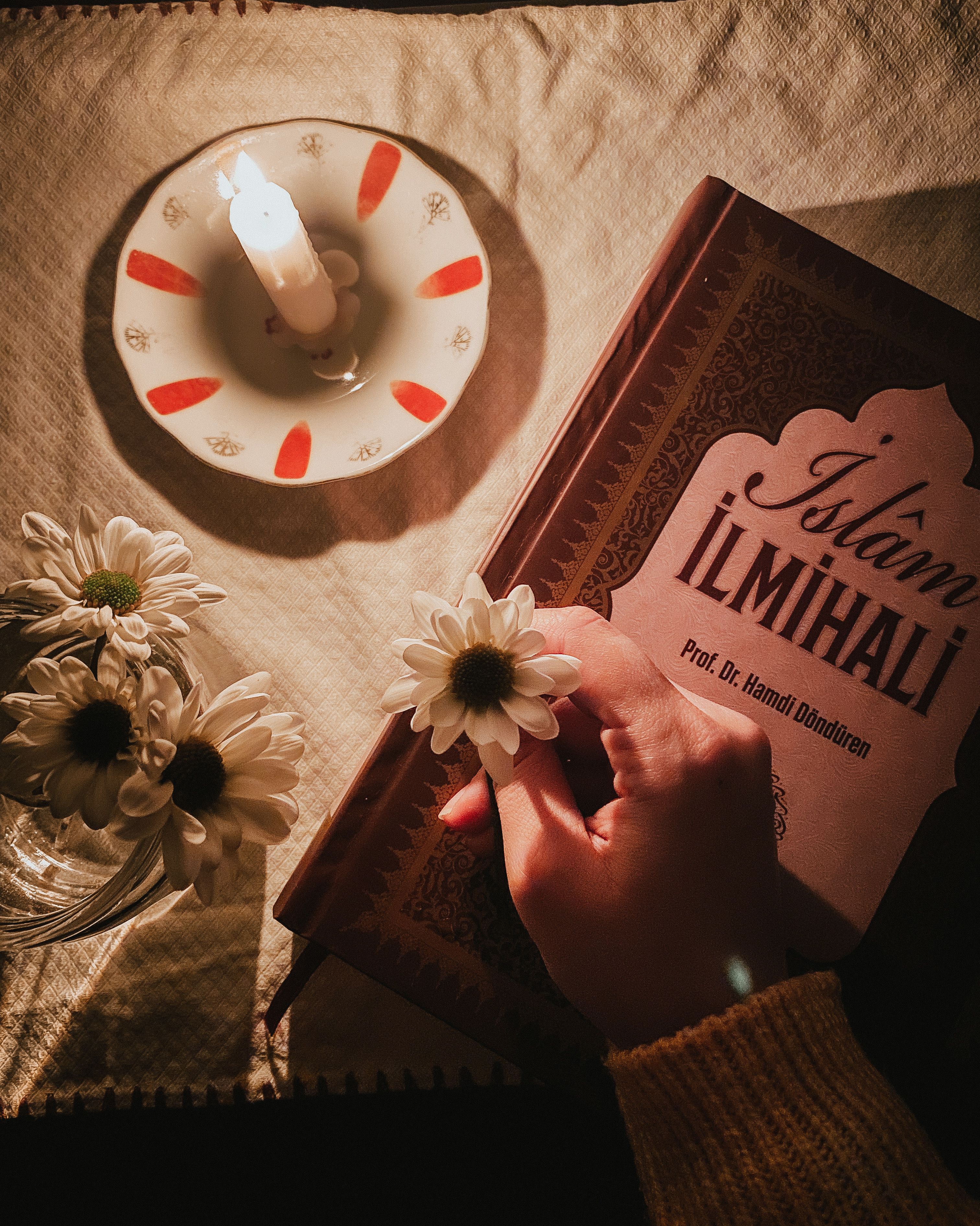 A hand holds a book next to a burning candle and flowers in a cozy evening setting.