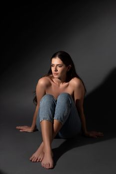 Portrait of a woman sitting barefoot in jeans, shadowy background enhancing mood.