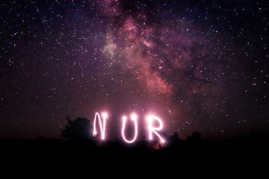 Night sky with stars and Milky Way galaxy featuring artistic light painting spelling 'NUR'.