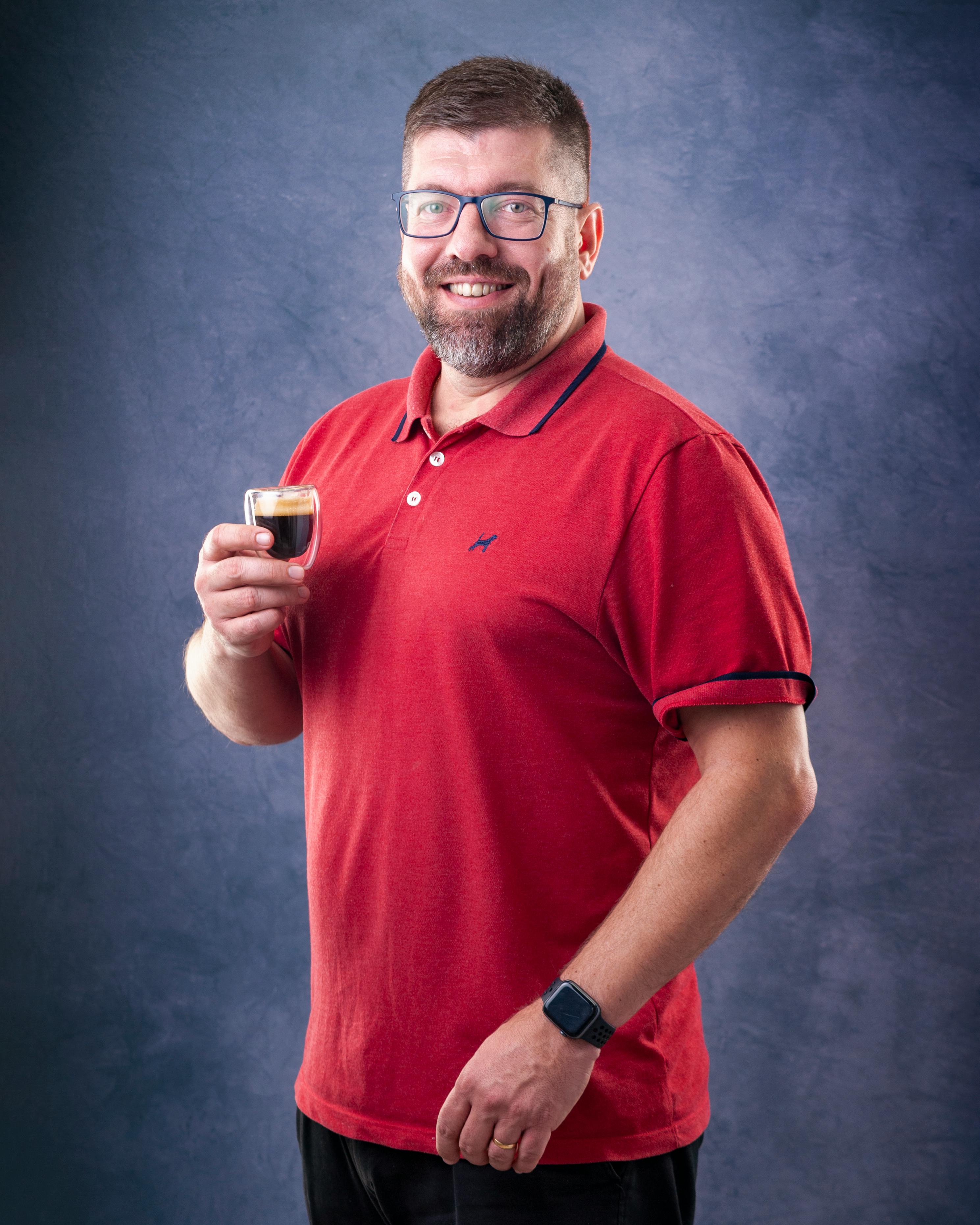 Portrait of a smiling man in glasses holding an espresso cup indoors.