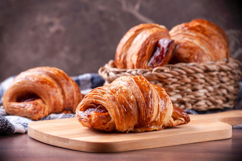 Golden, flaky croissants with visible layered lamination stacked on a cooling rack, one torn open to show the honeycomb interior.