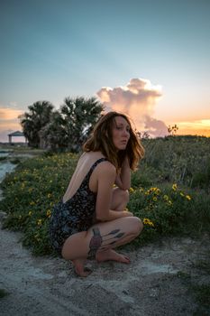 Woman crouching in a floral landscape at sunset, wearing a swimsuit, with a tattoo visible on her leg.