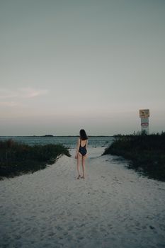 A woman in a bikini walking along a sandy beach path at dusk, with the sea in the background.