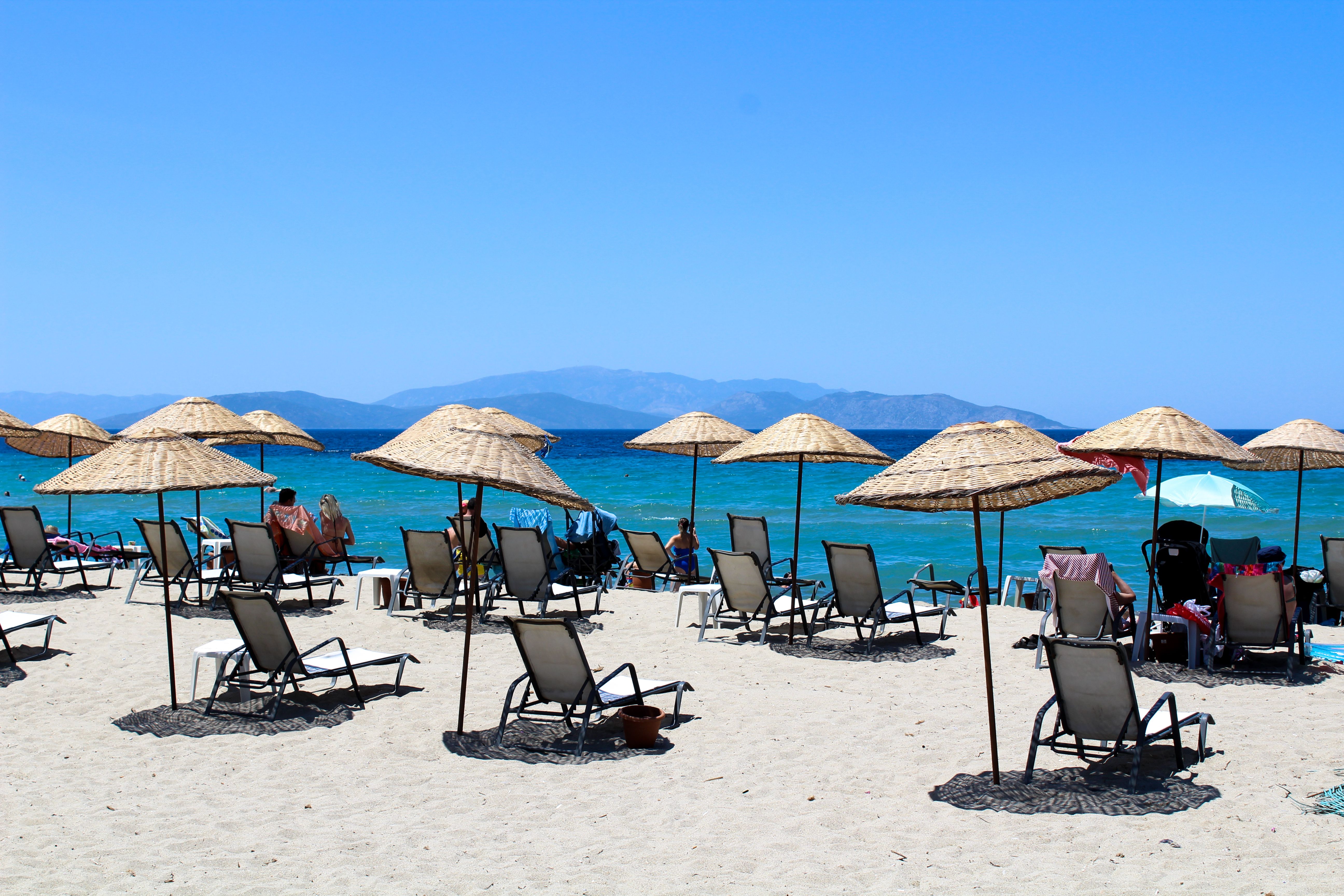 Turquoise waters of Bodrum beaches in Turkey with white sun loungers under blue umbrellas against backdrop of white-washed buildings and Aegean coastline