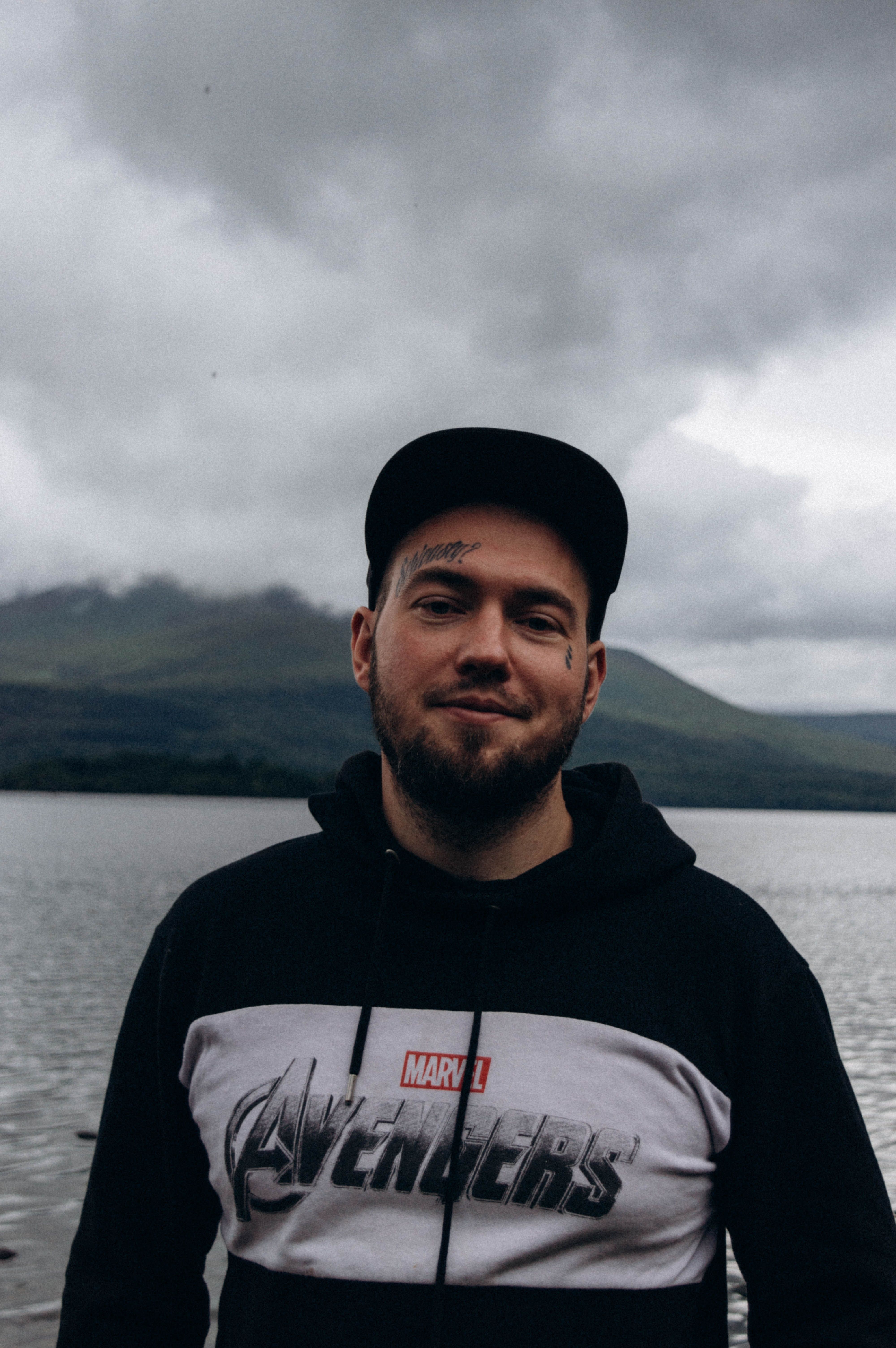 Bearded man in a cap and Marvel hoodie enjoying a serene lakeside view.