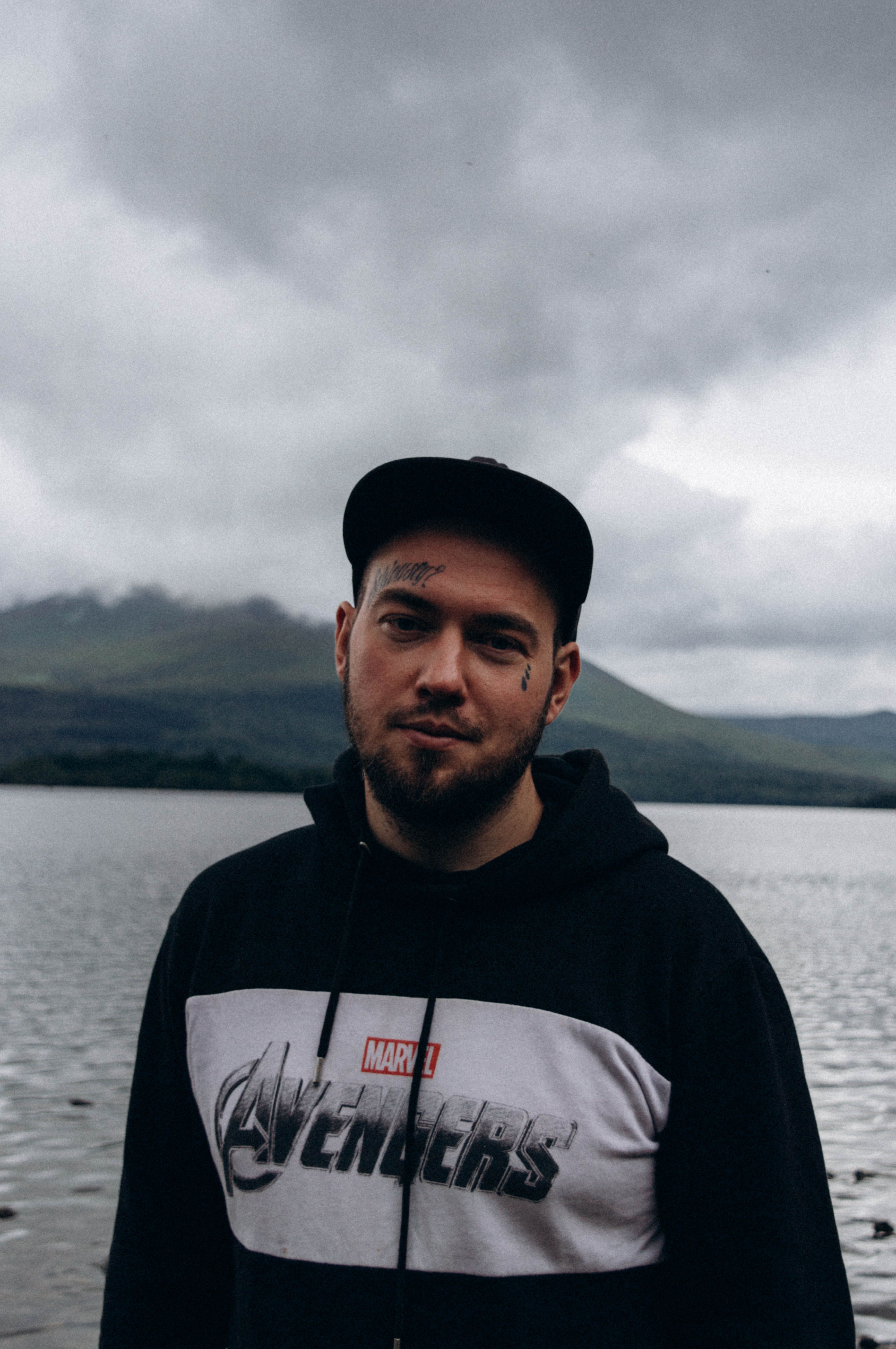 Man in black hoodie stands by a peaceful lake on a cloudy day.