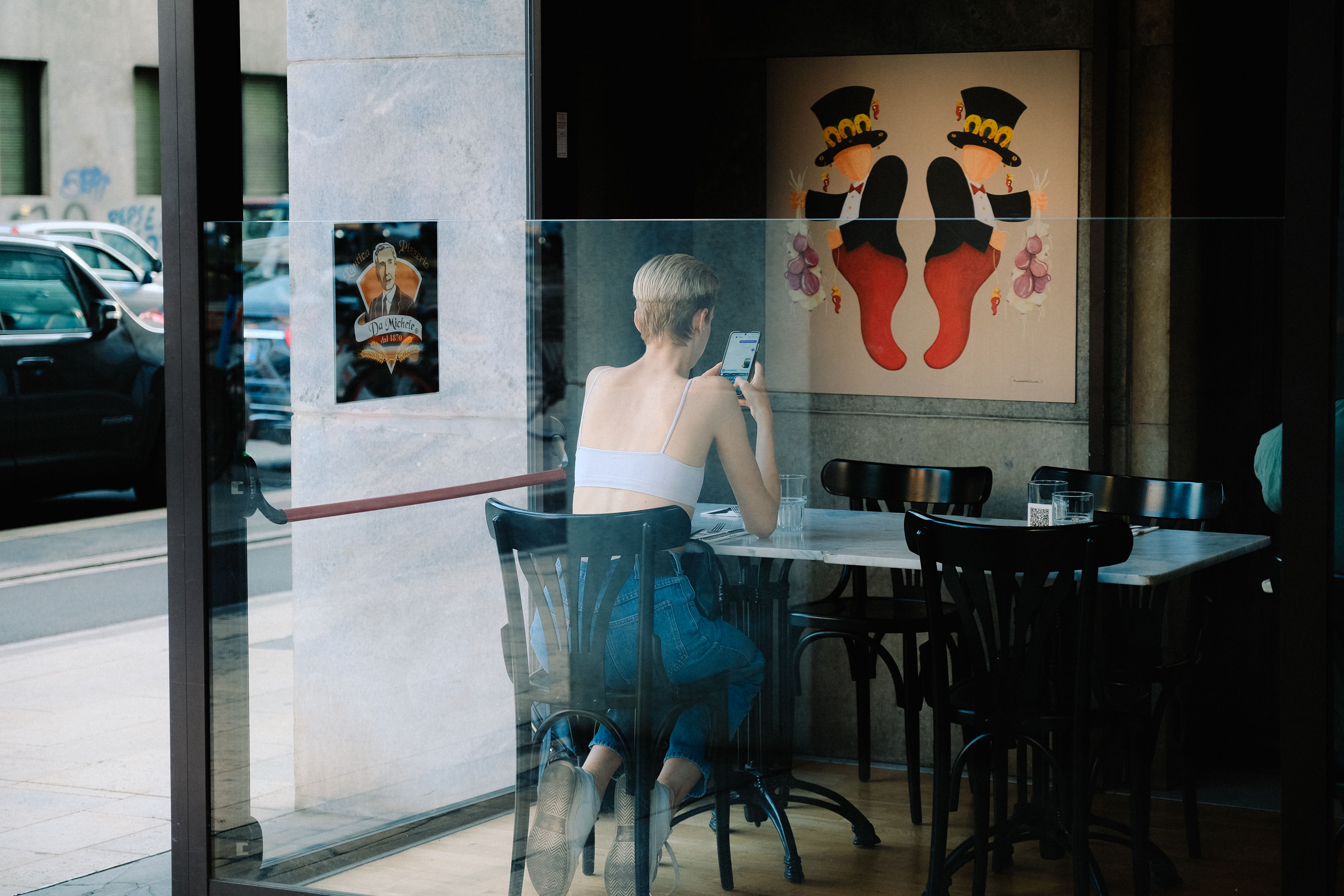 A woman sitting alone in a café, observed through glass — present but unacknowledged