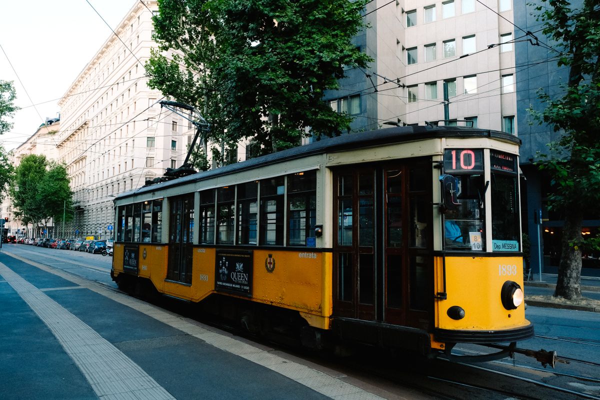 Tramvai galben clasic pe o stradă din Milano, cu clădiri istorice și copaci
