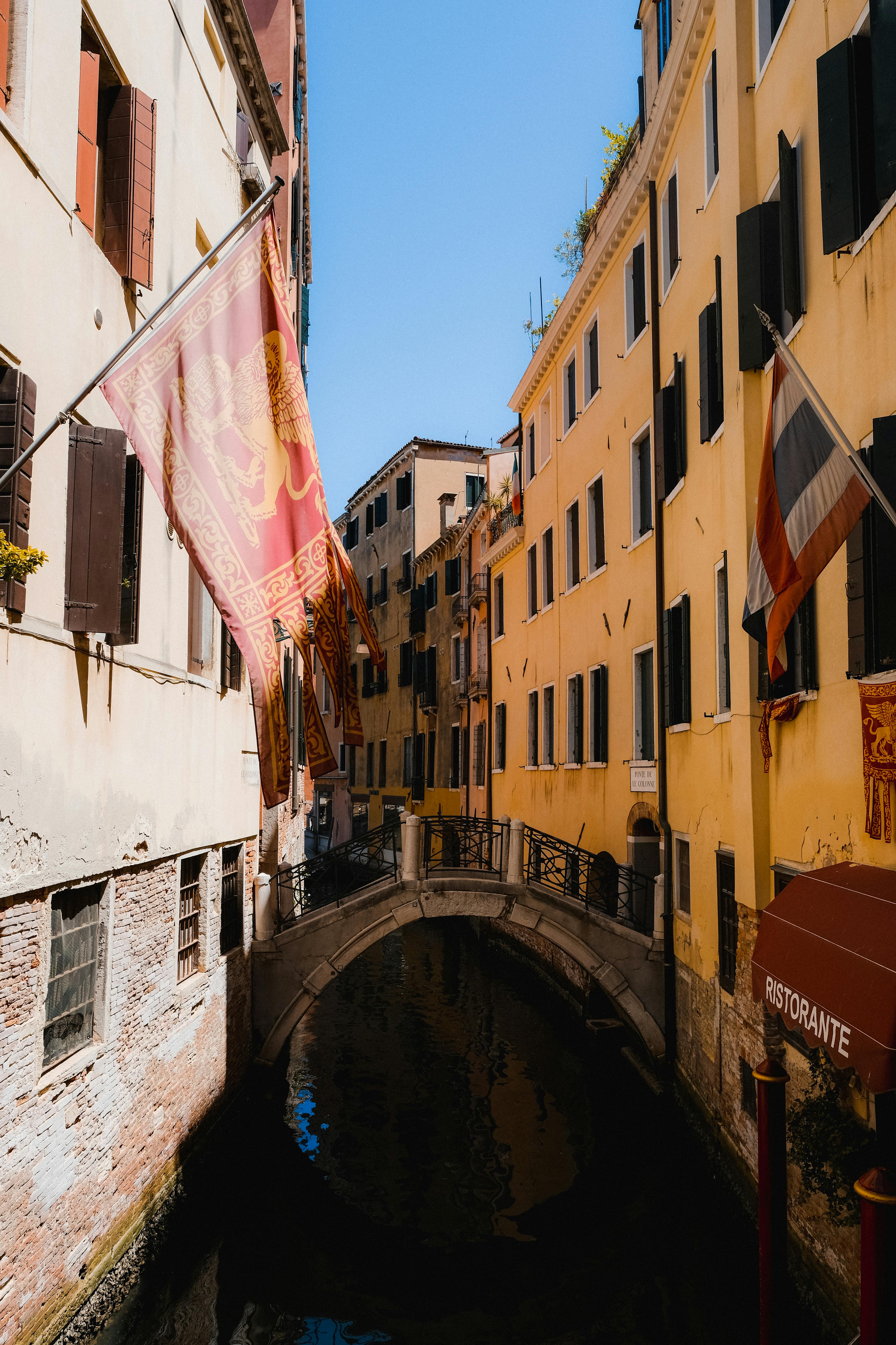 A picturesque Venice canal with traditional Venetian architecture and flags under a clear blue sky.