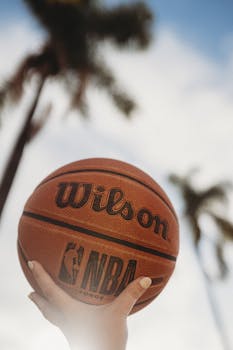 A hand holding a basketball with palm trees against a blue sky background.