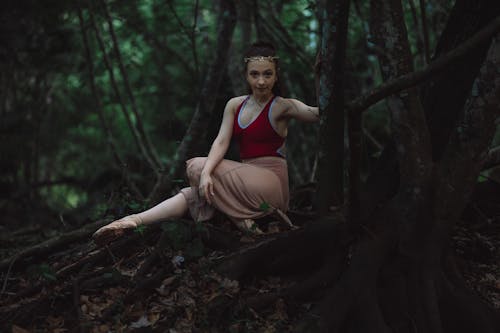 Woman Posing in Red Sleeveless Leotard Wearing Ballet Shoes Sitting on Tree Roots on a Forest