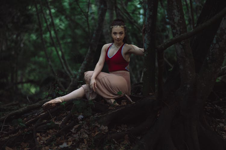 Woman Posing In Red Sleeveless Leotard Wearing Ballet Shoes Sitting On Tree Roots On A Forest