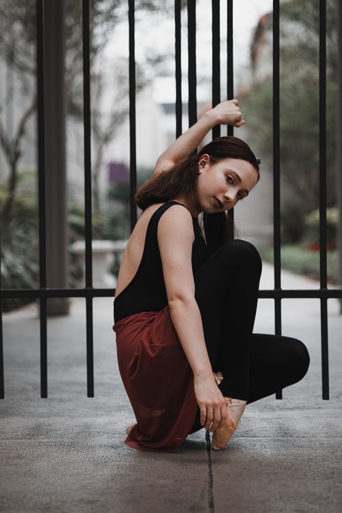 Free A graceful ballet dancer crouched and posing elegantly by a metal gate in an urban setting. Stock Photo