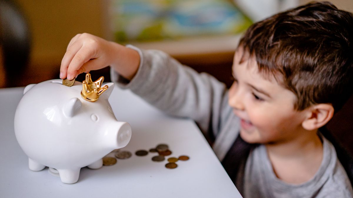 Menino sorrindo enquanto coloca moedas dentro de um cofrinho, aprendendo a poupar