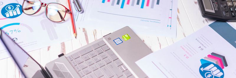 A cluttered desk with a laptop, financial reports, calculator, and glasses indicating a busy work environment.