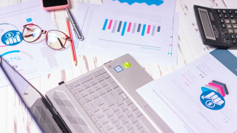 A cluttered desk with a laptop, financial reports, calculator, and glasses indicating a busy work environment.