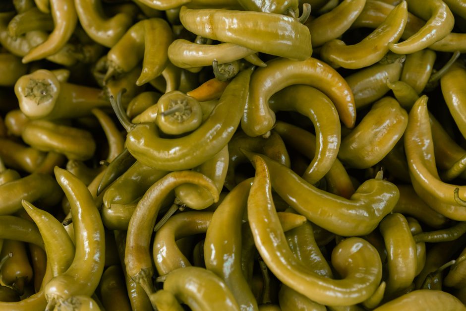 A glass jar of pale yellow-green pepperoncini peppers submerged in brine, with a few peppers scattered on a cutting board alongside their stems