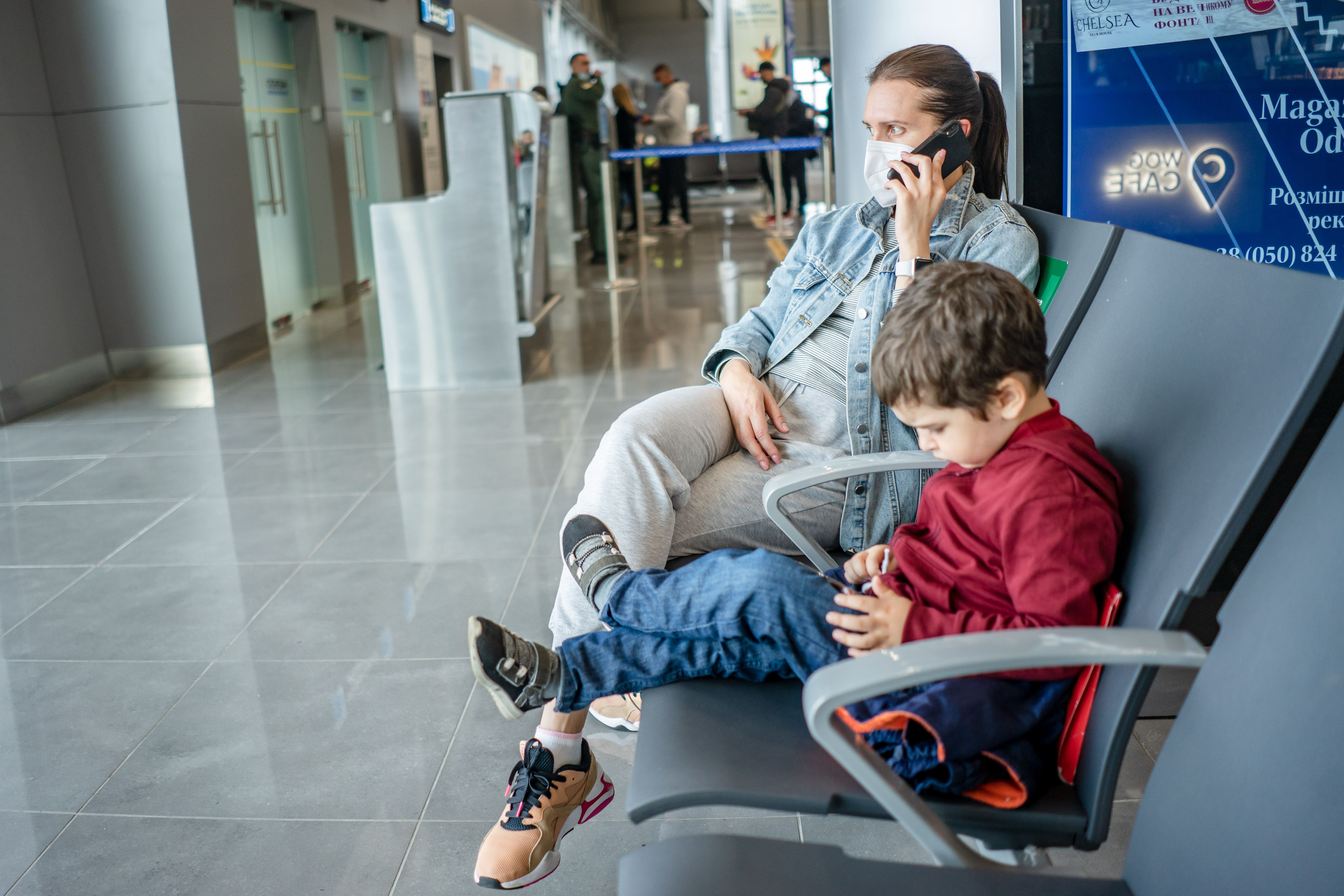 Mother wearing a face mask talks on her phone while her child waits beside her at an airport departure lounge