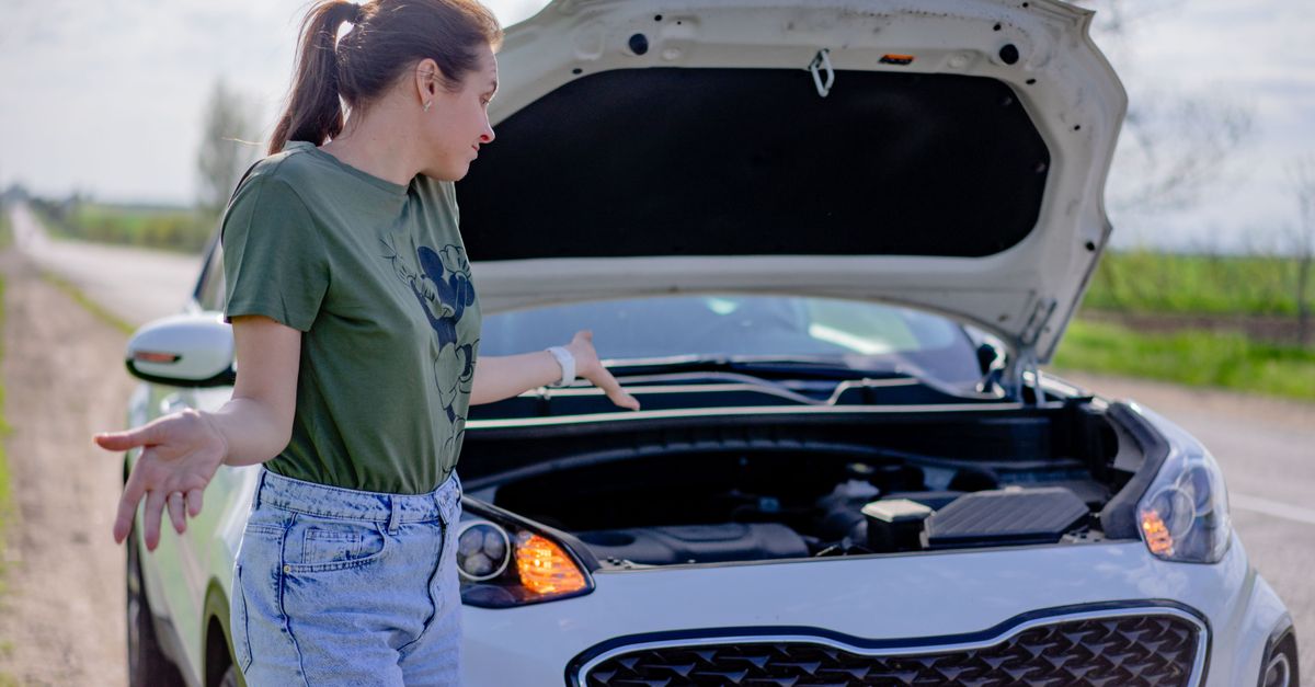 A woman looks puzzled by her broken car with an open hood on a rural road.