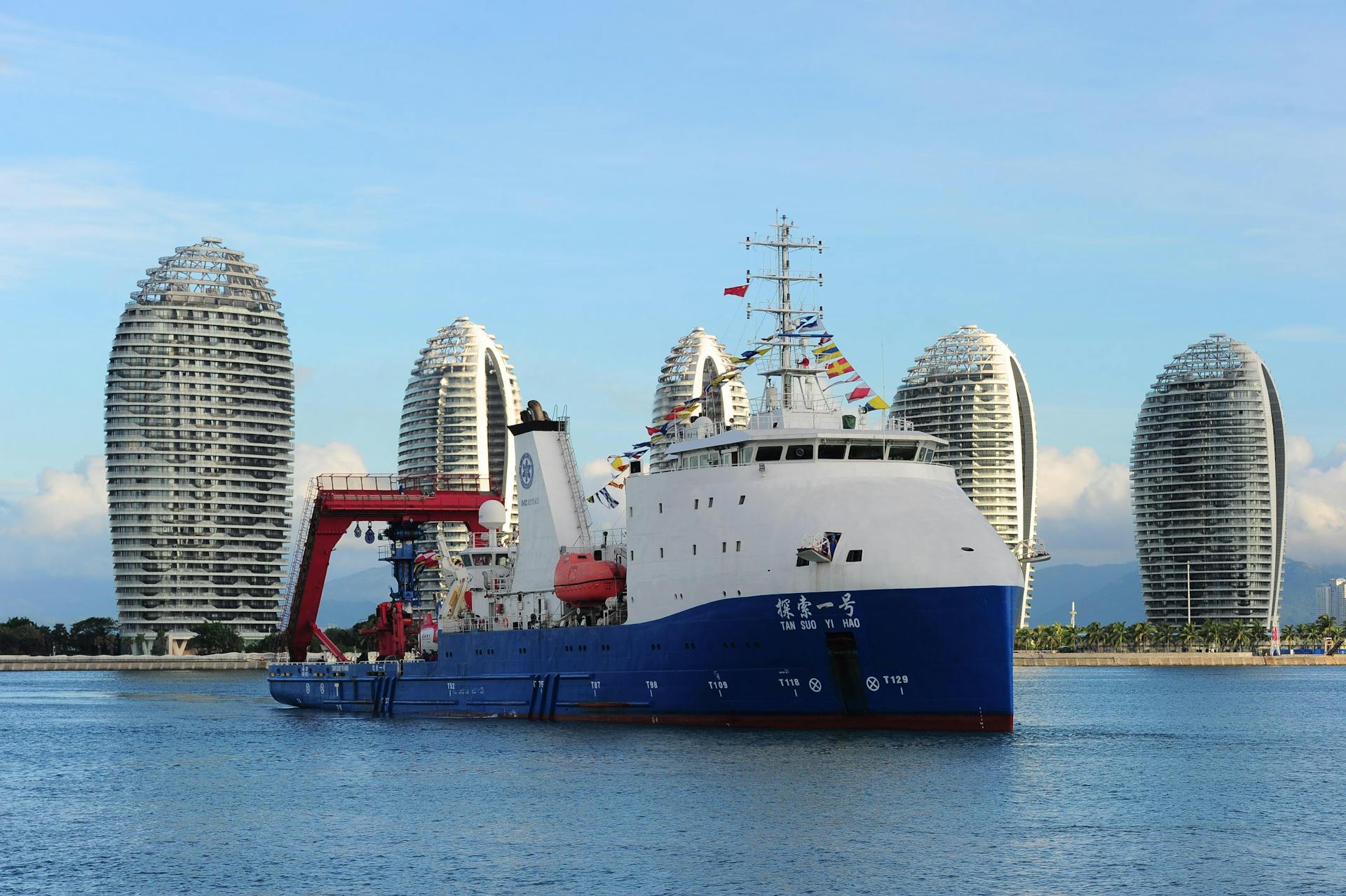 A modern cruise ship docked in the picturesque Sanya Bay, with iconic skyscrapers in the background.