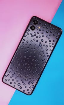 A sleek black smartphone covered in water droplets, placed on a vibrant pink and blue background, creates a striking visual contrast.