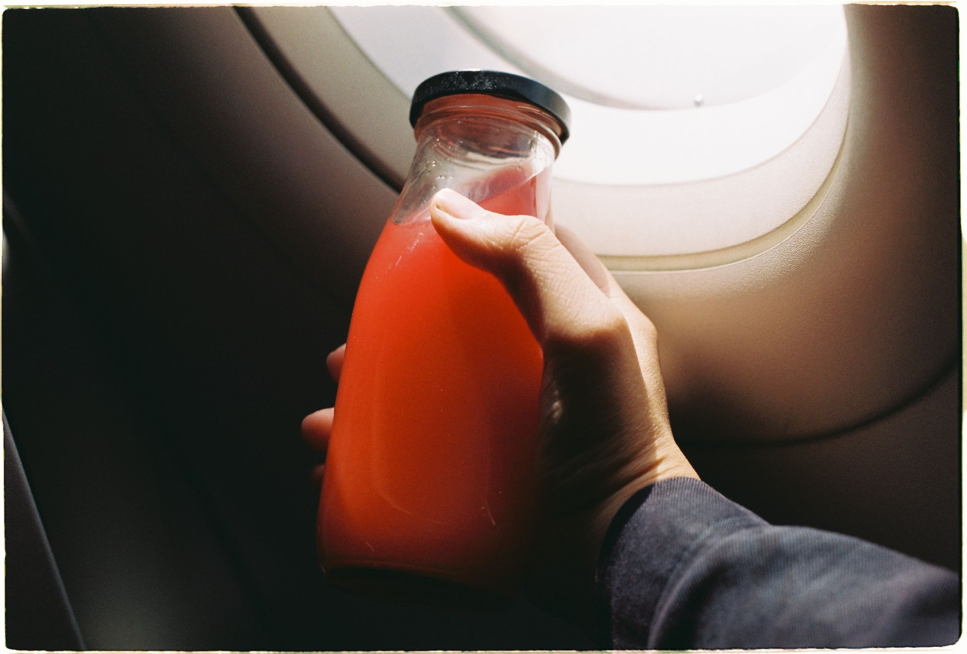 Hand holding a bottle of red juice next to an airplane window, natural light.