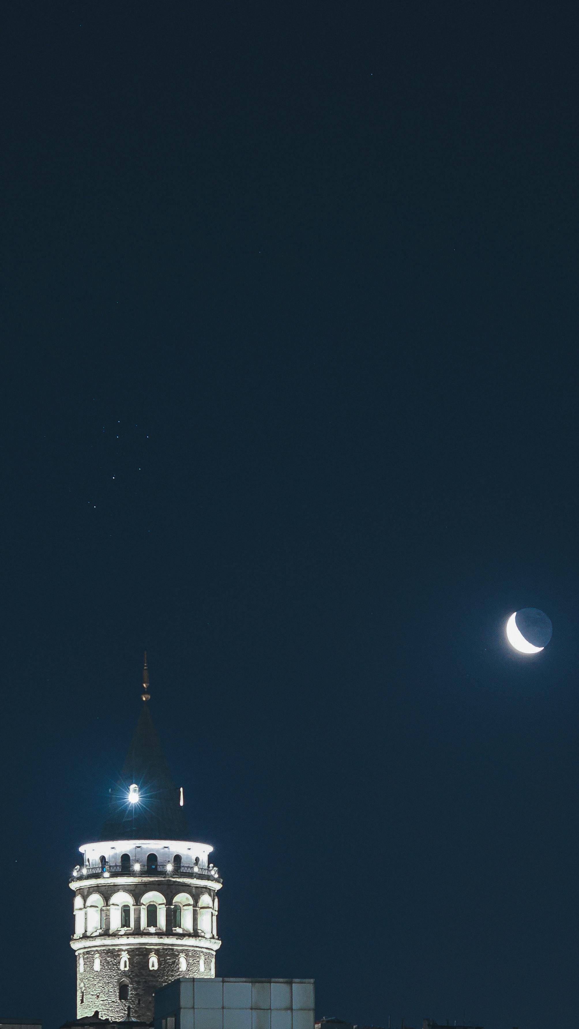 Illuminated Galata Tower with crescent moon against a night sky in İstanbul, Turkey.