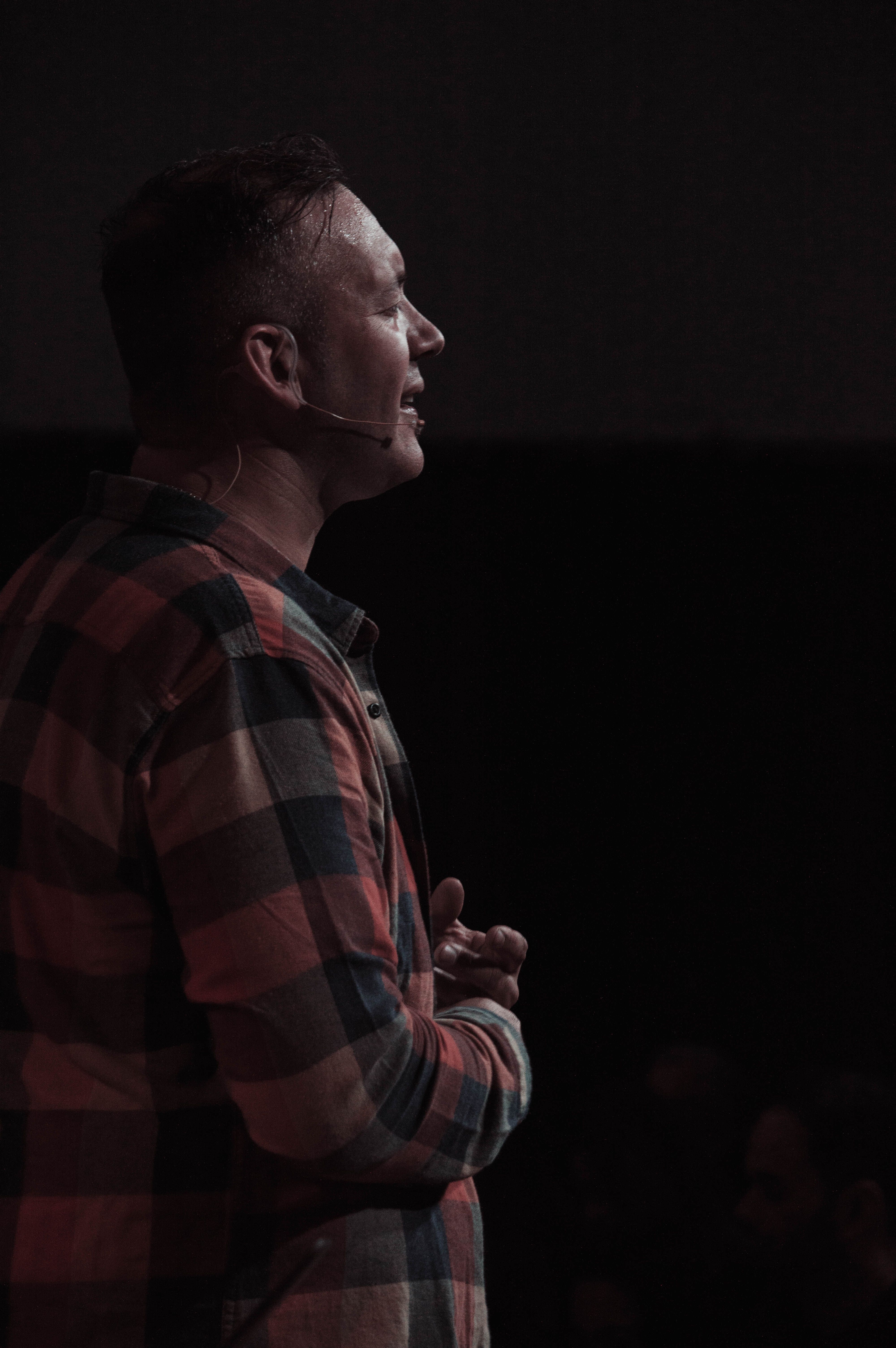 A man wearing a plaid shirt speaks into a headset microphone on a dimly lit stage.