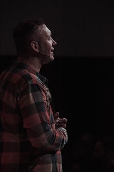 A man wearing a plaid shirt speaks into a headset microphone on a dimly lit stage.