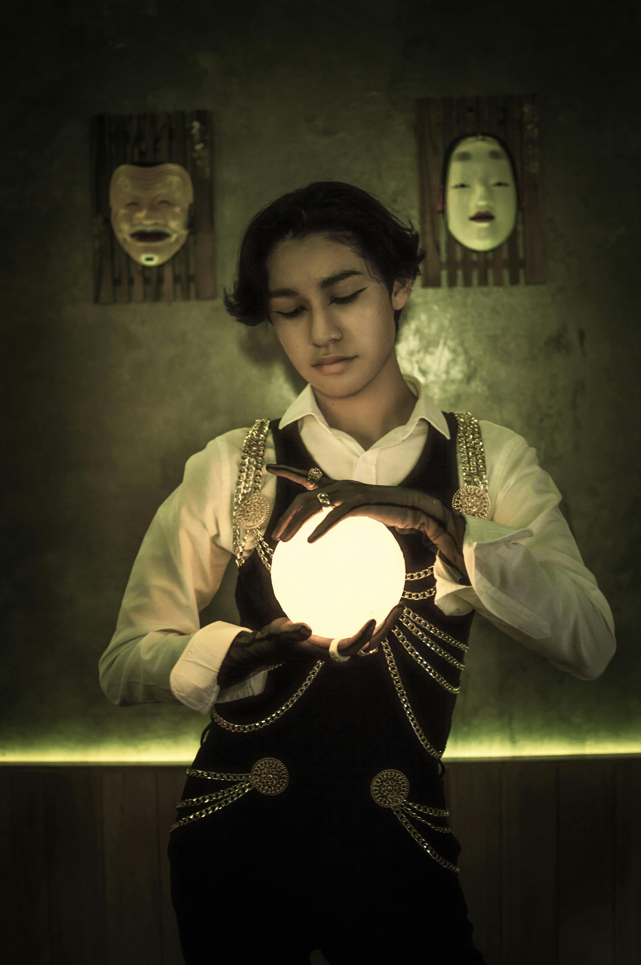 A fortune teller stands holding a glowing orb in a dimly lit room with masks on the wall.