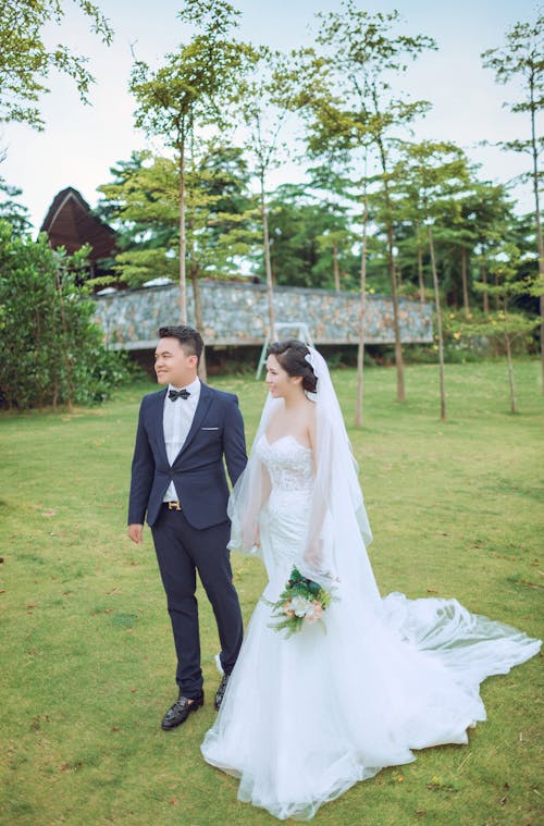 회색 건물 근처 웨딩 정장과 드레스를 입고 서있는 커플