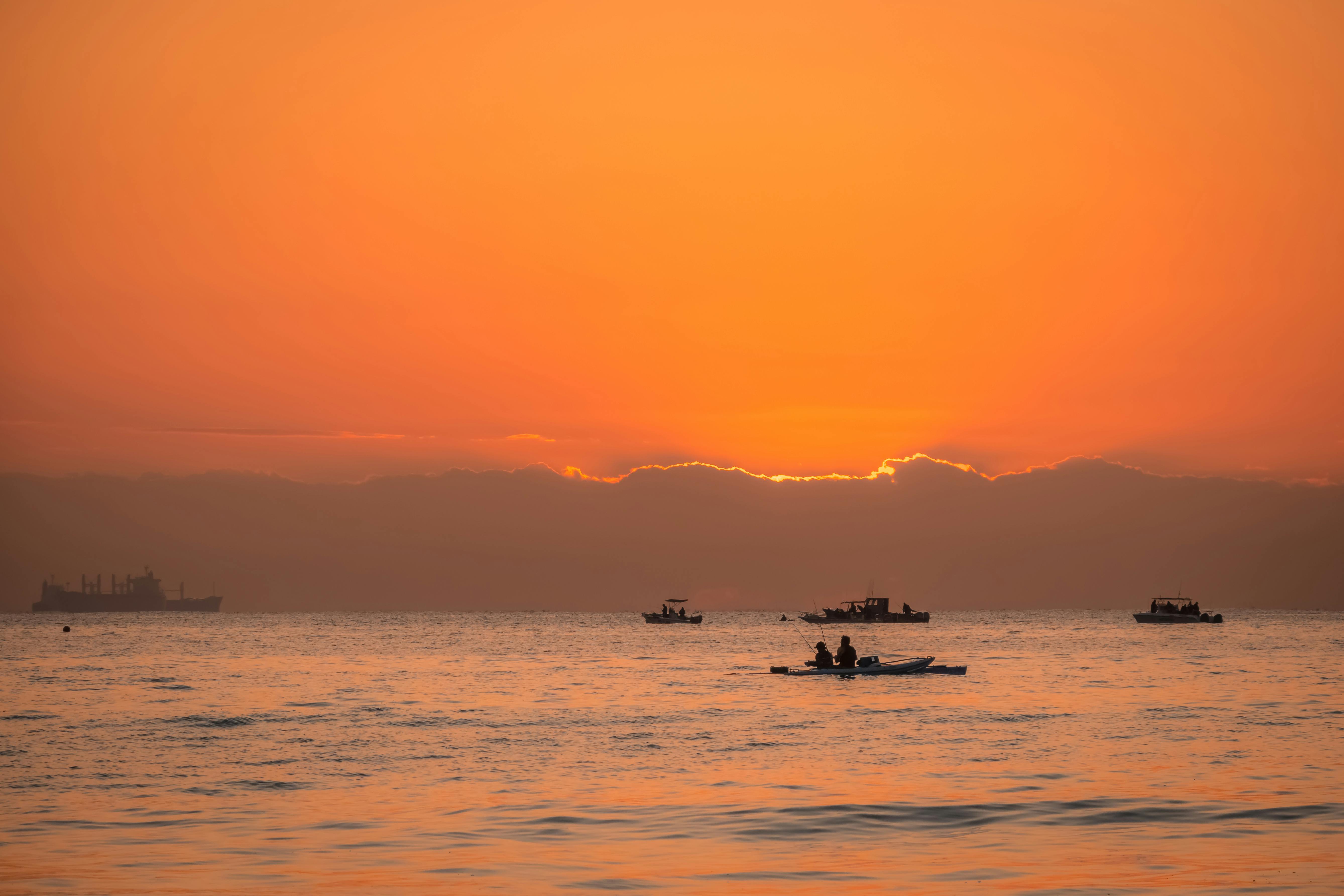 Silhouettes of boats and people at sunset with a vibrant orange sky and calm sea.