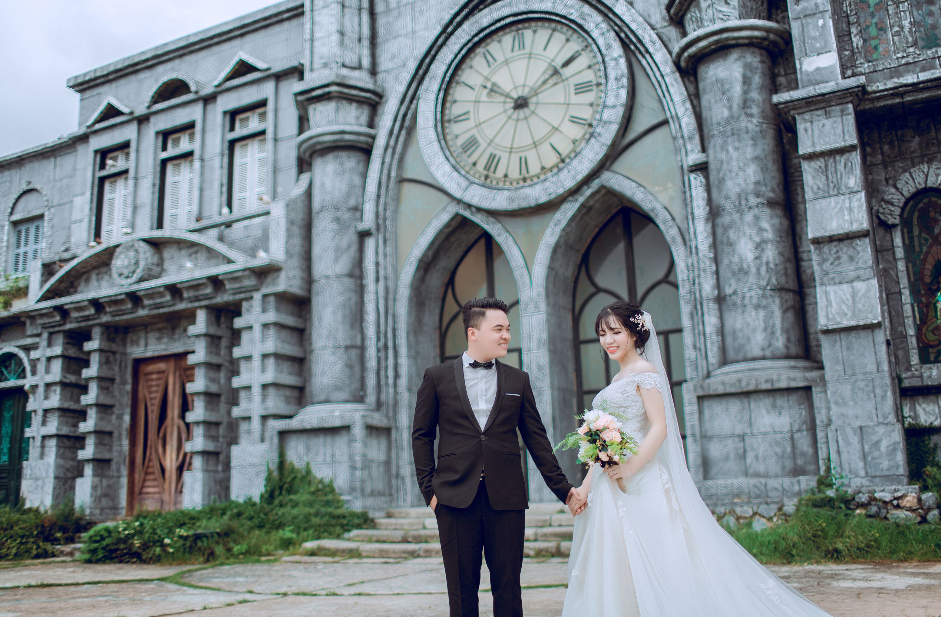 Elegant wedding couple posing outside a gothic-style building with a large clock and stone facade.