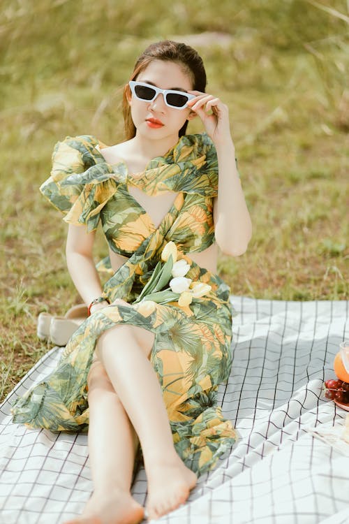 A Woman in a Green and Yellow Floral Dress Wearing White Framed Sunglasses Sitting on Green Grass