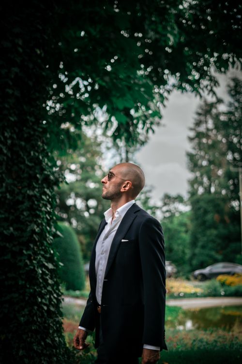 Free A stylish man in a black suit stands outdoors, surrounded by lush greenery and natural beauty. Stock Photo