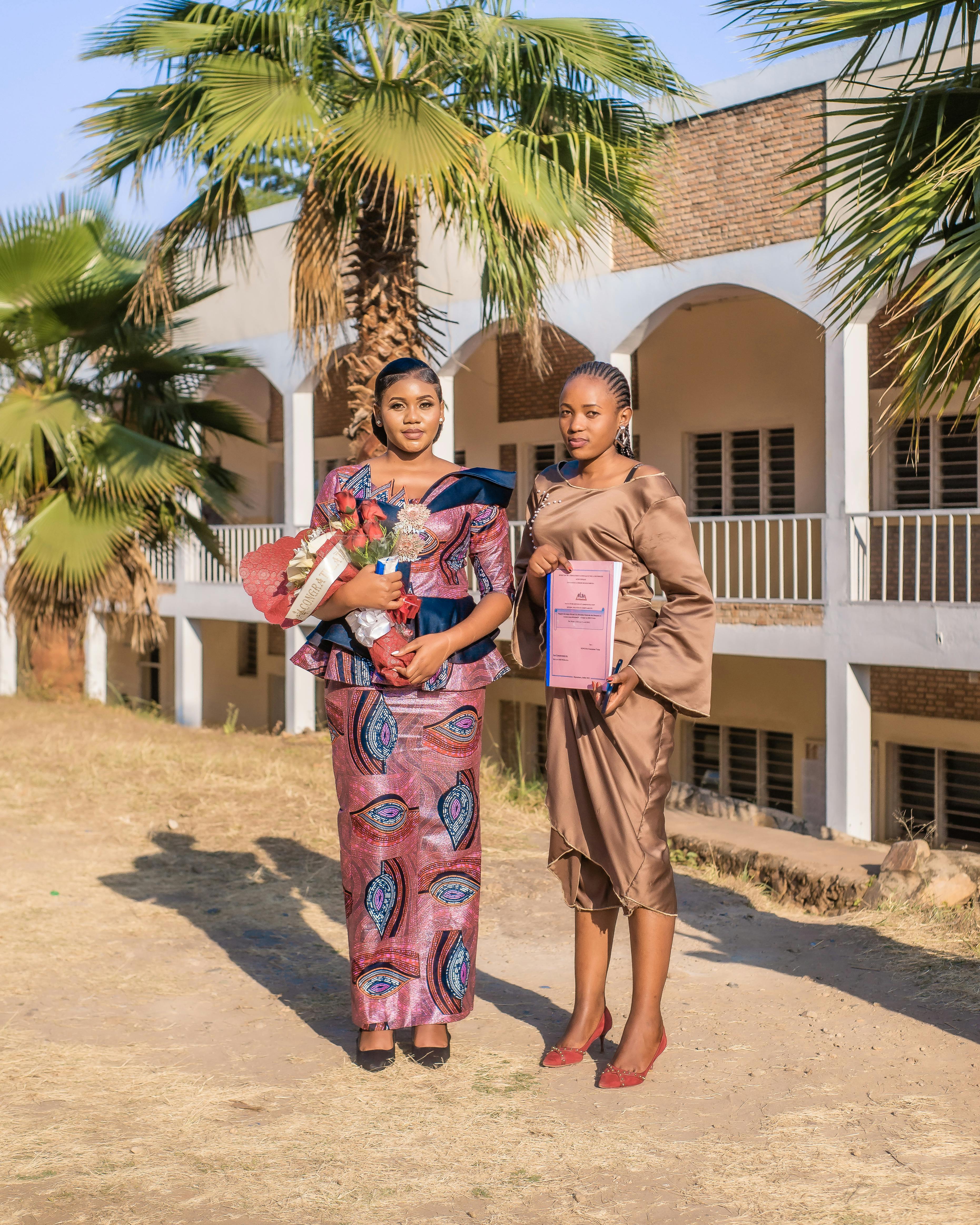 Two women celebrate graduation outdoors, holding bouquets and diplomas under palm trees.